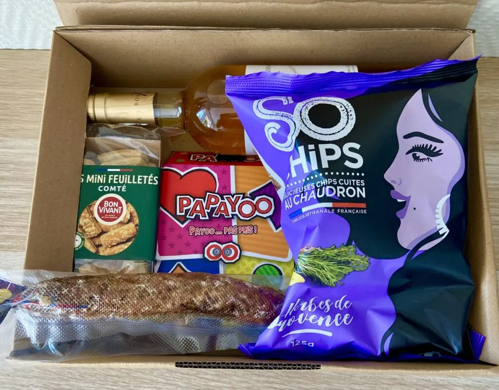 Une box Apero Game ouverte : une bouteille de vin, des mini feuilletés au comté, le jeu Papayoo, un saucisson et des chips So Chips aux herbes de Provence