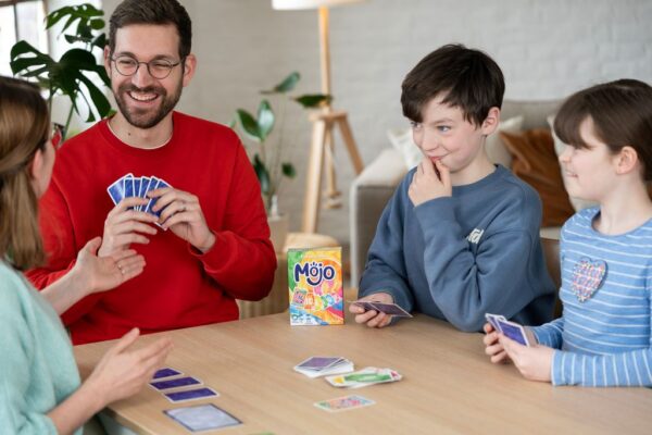 Photo d'une famille de 4, 2 adultes et 2 enfants, qui sourient en jouant sur une table au jeu de société Mojo